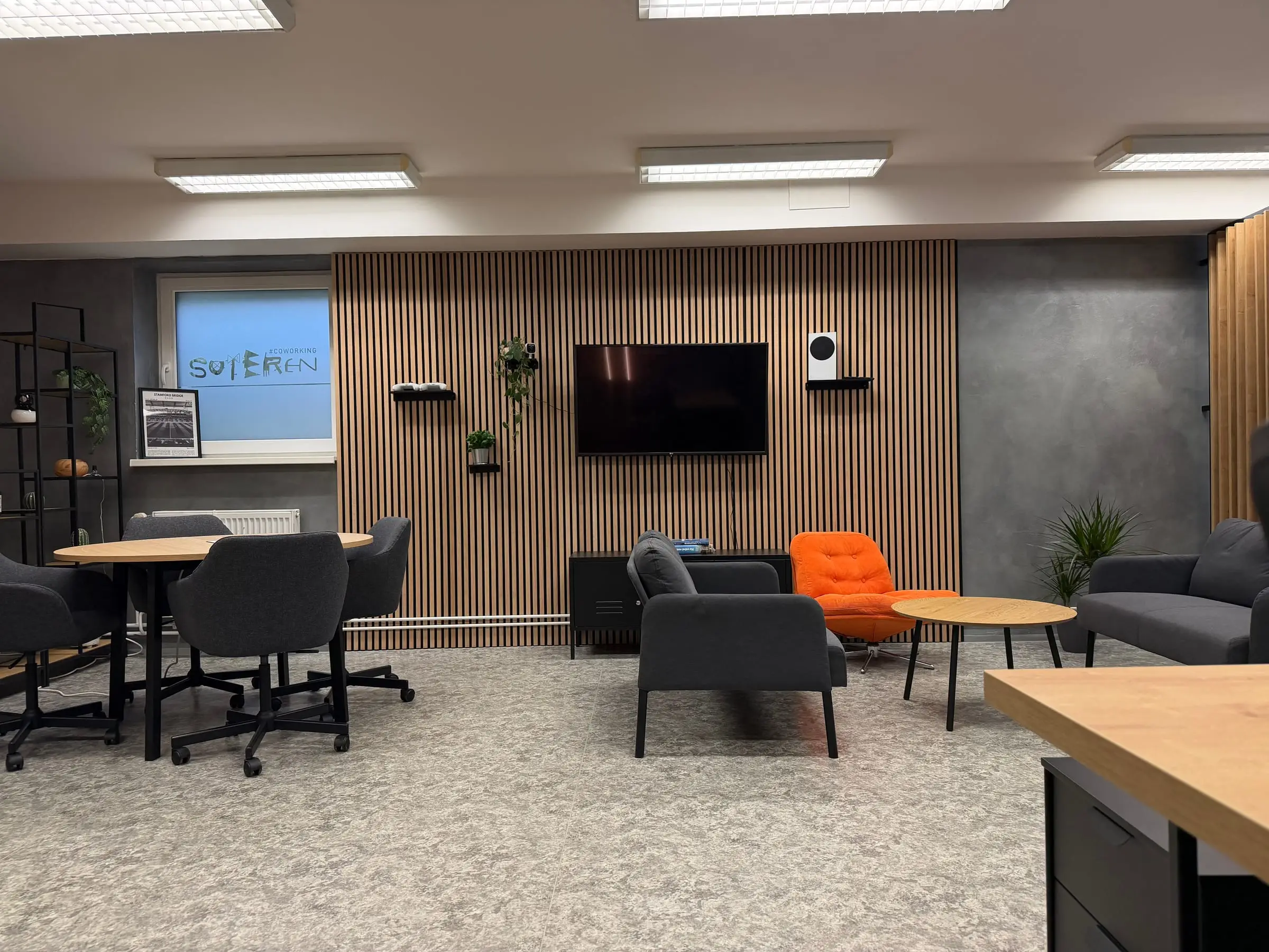 Coworking Suterén: kulatý stůl u okna s logem, relax zóna s oranžovým křeslem a lamelová stěna